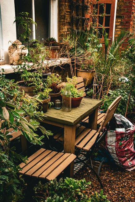 A cozy garden patio area adjacent to a brick house features a weathered wooden dining table and matching benches surrounded by lush, diverse potted plants with varying textures and shades of green, in